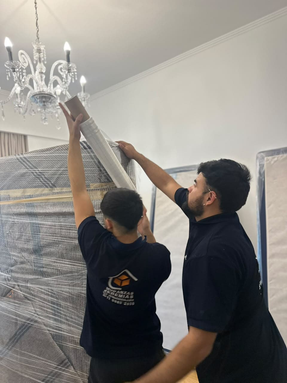 Dos integrantes del equipo envolviendo con film un mueble en un living con araña de luces